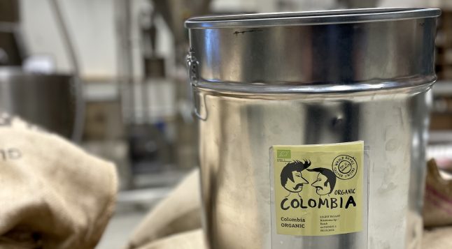 Meie mahemärgisega kohv – Colombia Magdalena, Sierra Nevada ORGANIC. Metallist kohvitünn, millel on kollane silt ja EL-i mahelogo, asetatud kohvikottide peale röstikojas.